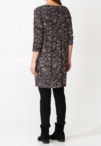 Kvinna som bär en tunika med svart och vitt paisleymönster, trekvartsärmar, svarta byxor och svarta mockaboots, stående med ryggen vänd.