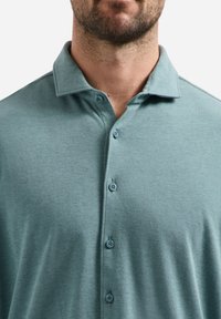 Man in een teal knopenoverhemd met kraag, die zijn nek, baard en bovenborst zichtbaar heeft tegen een effen achtergrond.