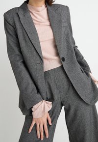 Šedý kostkovaný vlněný blazer s klopami, zapínáním na jeden knoflík a předními kapsami, v kombinaci s světle růžovým texturovaným topem.