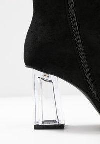 Botín de ante negro con cierre de cremallera y tacón bloque transparente, con un acabado brillante y una base cuadrada.