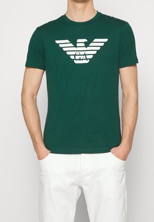 Homme portant un t-shirt à manches courtes vert foncé avec un logo d'aigle stylisé blanc et un pantalon blanc, les mains dans les poches.