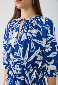 Robe bleue à motifs floraux avec des feuilles blanches, décolleté noué et taille élastique. Présente des manches bouffantes et une coupe décontractée, fabriquée en tissu léger.