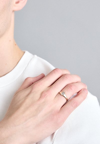 Zelts gredzens ar noapaļotu dizainu un mazu centrālo akmeni, uz rokas ar gaišu ādas toni, uz pelēka fona.