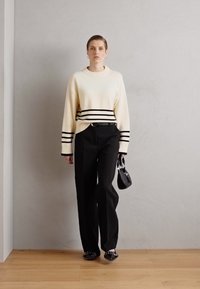 Crème trui met zwarte strepen, zwarte hoge taille broek, houdt een kleine zwarte tas vast. Glad textuur en een aangesloten silhouet.
