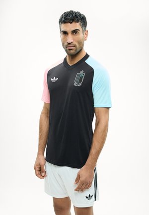 Άνδρας που φοράει μαύρη αθλητική φανέλα με ροζ και μπλε μανίκια, λευκό σορτς, και στέκεται μπροστά σε απλό λευκό φόντο.