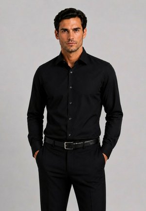 Homme debout, les mains dans les poches, portant une chemise noire cintrée et un pantalon noir devant un fond clair uni.