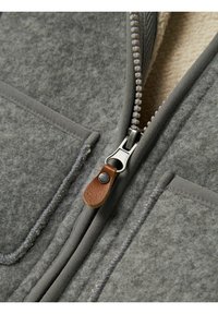 Giacca a zip grigia con una cerniera prominente dotata di un tirante in pelle, cuciture a contrasto e tasche laterali per riporre oggetti.