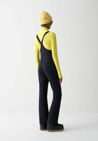 Persona che indossa pantaloni da neve neri, una maglietta a maniche lunghe gialla, un berretto giallo a fantasia, stivali neri e occhiali neri, in piedi davanti a uno sfondo bianco.