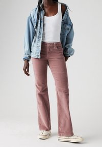 Veste en denim bleu clair, débardeur blanc et pantalons en velours côtelé rose évasés. Tissu texturé avec poches avant. Des baskets blanches complètent la tenue.