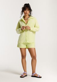 Licht limoen groene button-up shirt met lange mouwen, gecombineerd met bijpassende shorts. Schoeisel bestaat uit zwarte sandalen met paarse banden.