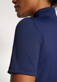 Námořnická modrá texturovaná polokošile s vysokým límcem, krátkými rukávy a plochými švy, zdůrazňující elegantní a sportovní design.