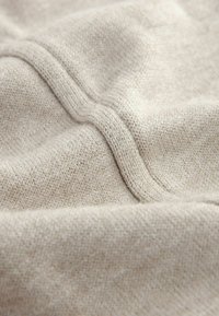 Beige Stoff mit einer weichen Textur und einem sichtbaren Strickmuster. Zwei parallele Nähte verlaufen über die Oberfläche und betonen die Weichheit des Materials.