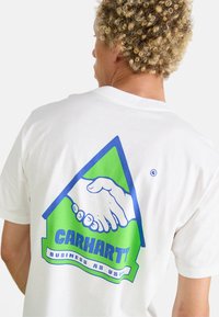 White cotton t-shirt featuring a large blue and green graphic on the back, depicting a handshake within a triangle, with "CARHARTT" text.