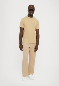 Beige t-shirt med en liten logotyp på bröstet, i kombination med ljusbeiga vida byxor och vita sneakers. Mörkbrun keps på huvudet.