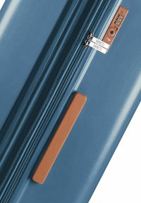 Valigia rigida blu con finitura liscia, dotata di doppie cerniere, un'etichetta per bagagli marrone e un pannello con lucchetto a combinazione sul lato.