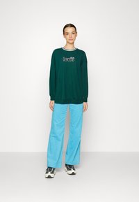 Green sweatshirt with ribbed neckline, featuring a graphic logo. Paired with light blue wide-leg pants and athletic shoes. Smooth fabric texture.