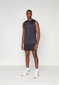 Svart ermeløs treningstopp med oransje logo, kombinert med svarte shorts. Modellen har på seg hvite joggesko med svarte detaljer og hvite sokker.