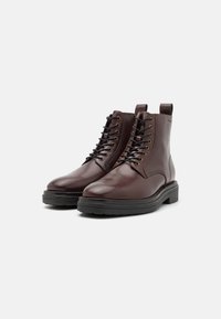 Bottines en cuir marron avec bouts ronds, semelles en caoutchouc noir et œillets en métal pour les lacets. Dispose d'une languette à l'arrière.