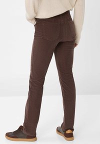 Persona che indossa pantaloni di velluto a coste marroni e sneakers marroni, con un maglione beige, in piedi davanti a uno sfondo chiaro e uniforme.