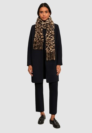 Cappotto navy con una silhouette dritta, indossato sopra una maglietta e pantaloni neri. Presenta una sciarpa leopardata beige con frange, abbinata a scarpe nere.