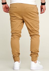 Khaki Jogginghosen mit elastischen Bündchen und Taschen auf der Rückseite. Getragen mit weißen Sneakers, die den strukturierten Stoff und die schmal zulaufende Passform zeigen.