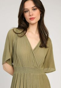 Robe vert olive avec un col en V, des manches courtes flottantes et un tissu texturé. Comprend une taille smockée et des détails délicats le long du col.