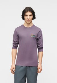 Νέος άνδρας με μεσαίου μήκους σκούρα μαλλιά, φορώντας μωβ πουκάμισο Billabong με μακριά μανίκια και μπλε-γκρι παντελόνι, στέκεται μπροστά σε απλό φόντο.