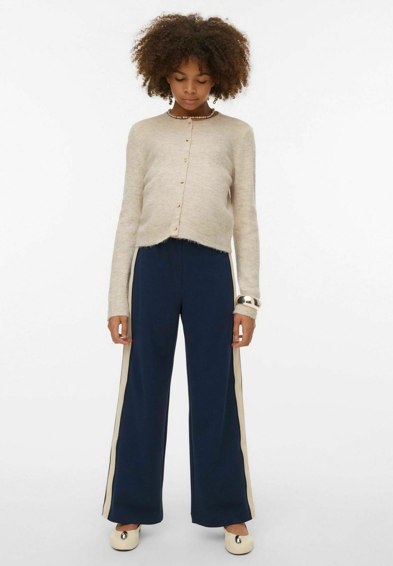 Béžový pletený kardigan na zapínán�í s knoflíky, doplněný tmavě modrými kalhotami se širokými nohavicemi a krémovými pruhy po stranách. Modelka má obuté boty s kulatou špičkou.