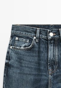 Dunkelblaue Jeans aus Denim mit vorderer Knopfleiste, Gürtelschlaufen und einer kleinen Münztasche oberhalb der rechten Vordertasche.