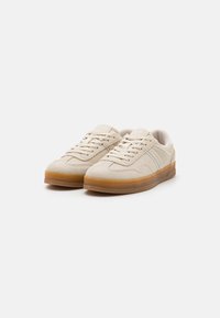 Beige suede sneakers with a textured surface, rounded toe, and gum rubber sole; features logo accents and lace-up design.