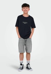 T-shirt noir avec l'imprimé "GALERIE PARIS", shorts en denim gris, baskets montantes noires et chaussettes blanches avec des rayures noires.