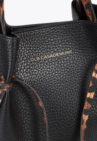 Schwarze Lederhandtasche mit strukturiertem Oberflächen und einem goldenen Logo "LOLA CASADEMUNT", mit Tierdruckakzenten an den Trägern.