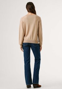 Bežna pletena jopica s širokim izrezom, dolgimi rokavi in rebrastim robom, v kombinaciji s temno modrimi razširjenimi kavbojkami in rjavimi čevlji na peto.