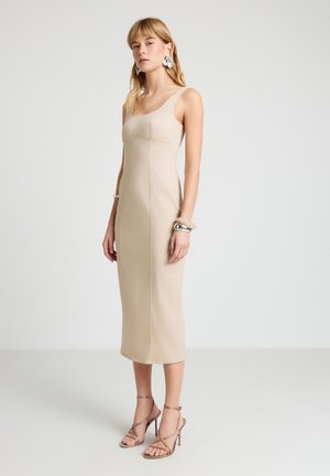 Donna che indossa un vestito midi beige senza maniche con silhouette aderente, abbinato a sandali con tacco alto con cinturini e gioielli argentati vistosi su sfondo bianco.