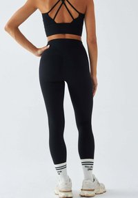 Czarne legginsy sportowe z wysokim stanem, w połączeniu z dopasowanym czarnym topem z krzyżującymi się paskami. Białe skarpetki i beżowe sneakersy.