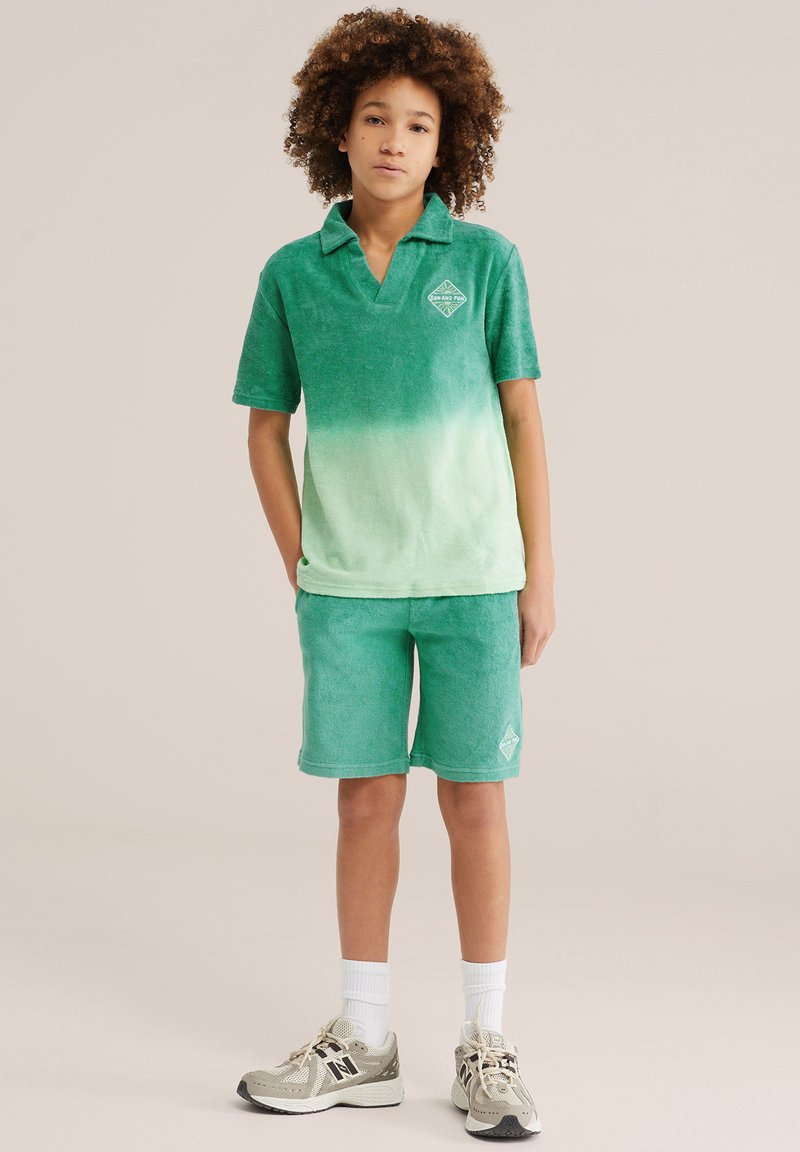 Enfant aux cheveux bouclés portant un polo dégradé vert et un short assorti, des chaussettes blanches et des baskets beiges, debout devant un fond uni.