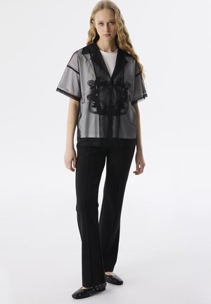 Camicia nera trasparente con bottoni e maniche corte, caratterizzata da un design ricamato; abbinata a pantaloni neri aderenti e scarpe nere flat.