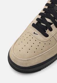 Beige zamšas snekeri ar melnajām šņorēm un akcentiem. Iespējas ietver perforācijas uz pirksta, gumijas zoli un mazu melnu logo sānos.