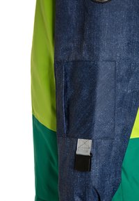 Primo piano di una manica in denim con tasca a toppe e clip nera, accanto a un pannello di giacca in tessuto verde e teal.