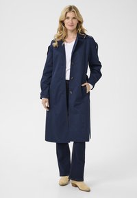 Marineblauwe trenchcoat van waterdicht materiaal, met een double-breasted ontwerp, knoopsluiting en zijzakken, gecombineerd met flared jeans.