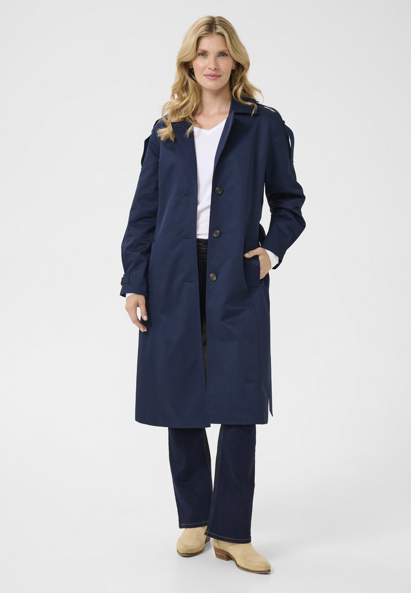 Marineblauwe trenchcoat van waterdicht materiaal, met een double-breasted ontwerp, knoopsluiting en zijzakken, gecombineerd met flared jeans.
