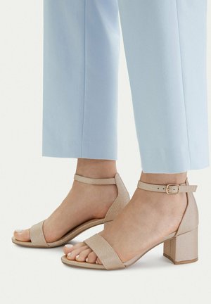 Füße in beigen Sandalen mit Blockabsatz und Knöchelriemen, kombiniert mit hellblauen, gekürzten Hosen vor weißem Hintergrund.