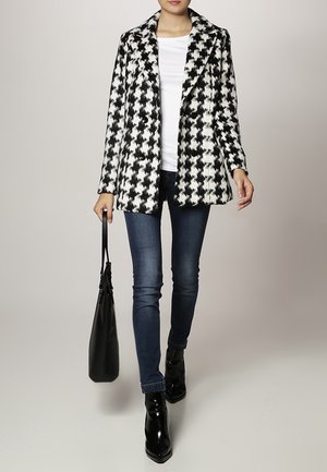 Cappotto a motivo pied de poule in bianco e nero con colletto, design doppiopetto, abbinato a jeans e borsa nera. Stivaletti lucidi alla caviglia.
