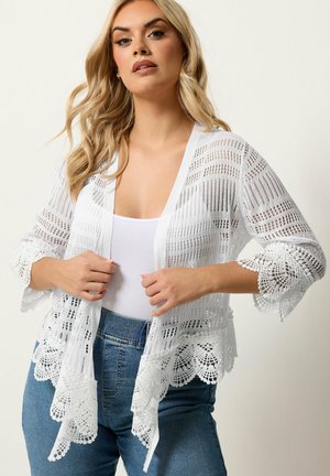 Kvinde med bølget blondt hår iført en hvid blondecardigan over en hvid top og blå jeans, justerer cardiganen foran en ensfarvet baggrund.