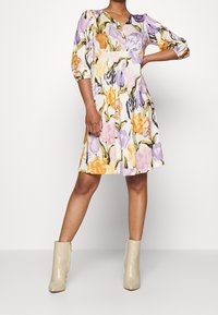 Blumenkleid mit lila, orange und gelben Mustern auf hellem Hintergrund; V-Ausschnitt, Puffärmel und plissierter Rock. Beigefarbene Knöchelstiefel.