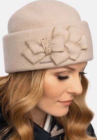 Sombrero de fieltro beige claro con un detalle de lazo tejido, acentuado por costuras doradas y gemas artificiales. Presenta una etiqueta de logo metálica. Textura suave.