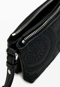 Sac à bandoulière en cuir noir avec motif circulaire embossé, compartiments à double fermeture éclair et accents en métal argenté. Texture lisse.
