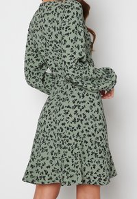 Green floral dress with long sleeves, textured fabric, black flower pattern, fitted waist, and flared hem.