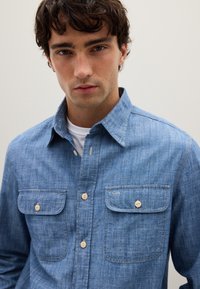 Camisa de denim azul claro con dos bolsillos en el pecho, diseño de botones y una tela texturizada. Cuenta con un collar y acentos de botones beige.