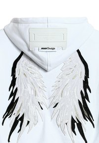 Sudadera con capucha blanca con grandes alas bordadas en negro y plata en la espalda, con tela texturizada y una etiqueta en el cuello.